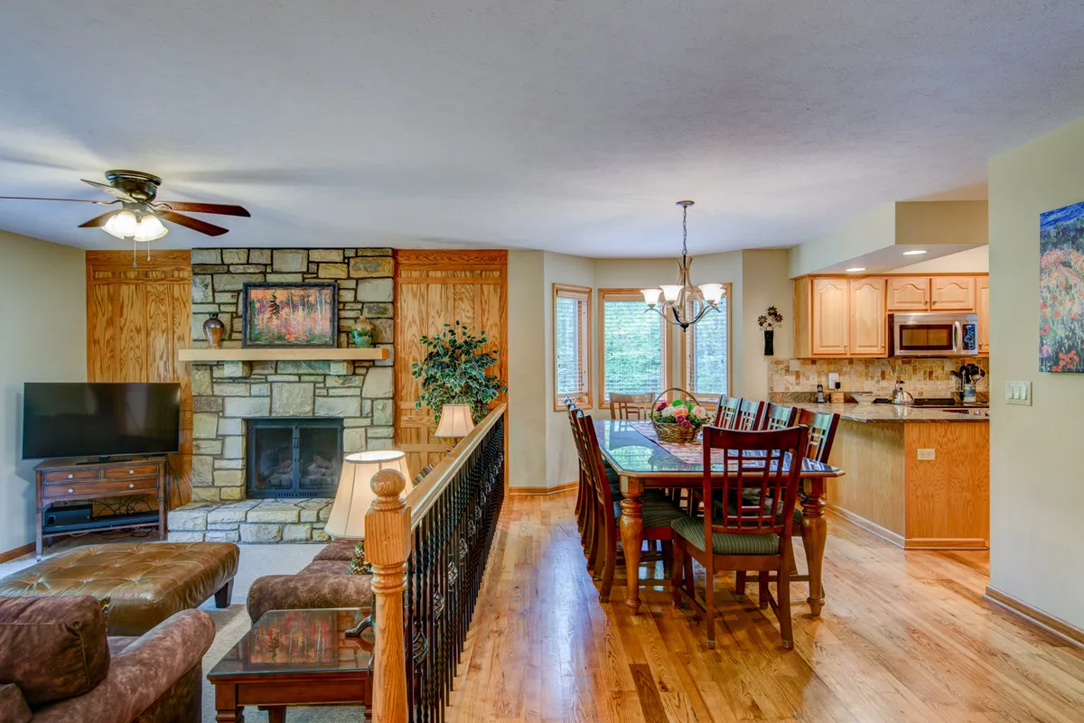 This image features a cozy interior living space with a fireplace, a dining table, a well-equipped kitchen area, and large windows. The room is sunlit and furnished for comfort, emphasizing both relaxation and togetherness.