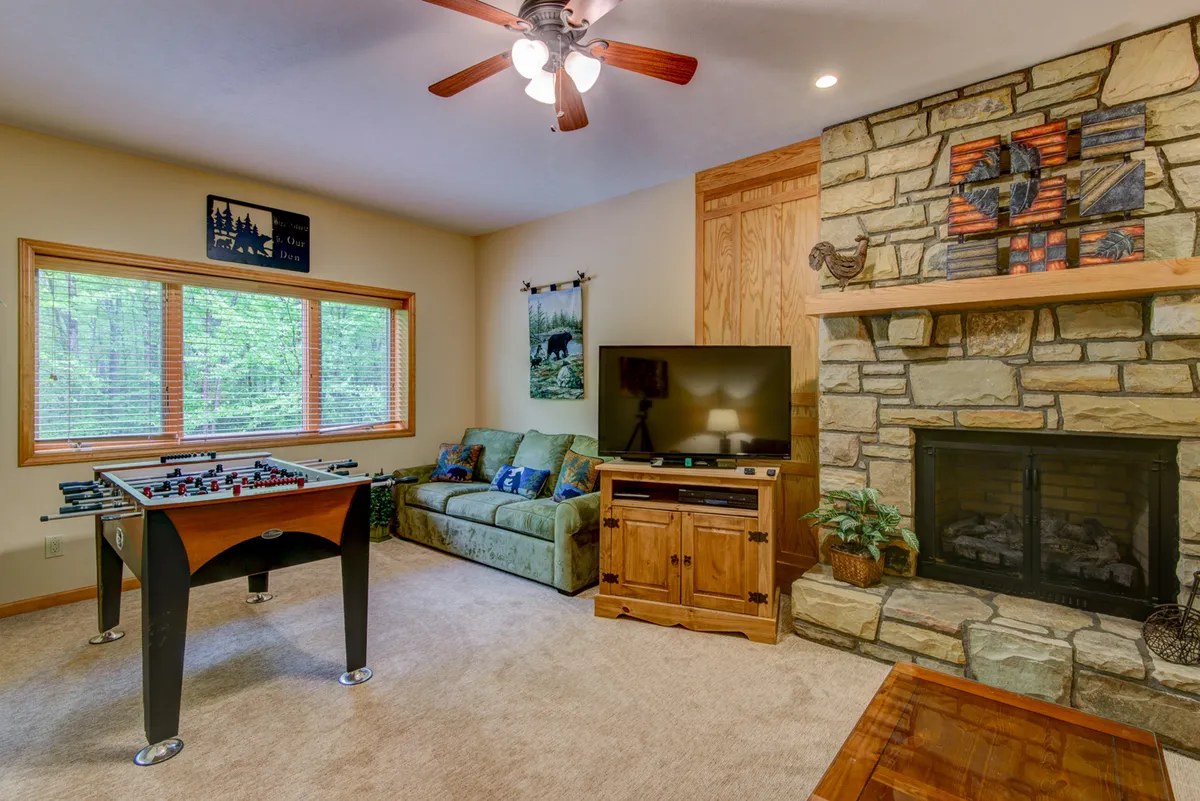 This interior image showcases the den's core entertainment features: a stone fireplace creates a cozy focal point, while a foosball table is positioned in the foreground, hinting at lively game nights. A comfortable sofa faces the television, and the overall layout supports both relaxation and play. Warm lighting and inviting furniture reinforce the room's function as a social and flexible retreat.