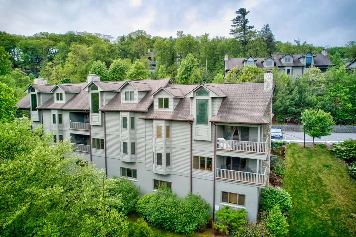 A multi-story condominium building set against lush greenery, with several balconies overlooking the landscape. People can be seen relaxing on balconies and walking in the tranquil garden below, suggesting a peaceful setting for group relaxation and connection with nature.