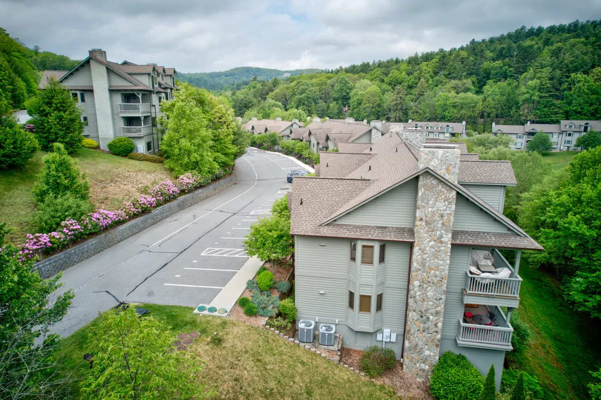 A wide shot of the apartment buildings nestled among beautifully landscaped gardens, with the parking area in the foreground and mountain views in the background. The image exudes an atmosphere of relaxed outdoor living, with people strolling and socializing in the green spaces.