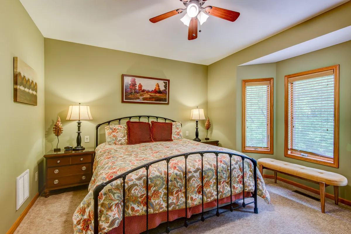 This image features a cozy bedroom with a bed dressed in floral bedding, nightstands with lamps on each side, and a window with blinds. Sunlight streams into the room, creating a warm and welcoming atmosphere.