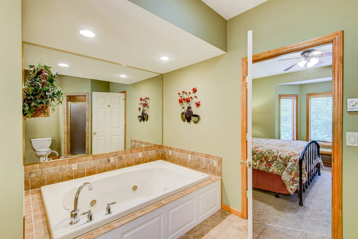 A tranquil bathroom scene showing a large bathtub, mirror, decorative plants, and a glimpse into an adjoining bedroom. The space feels spa-like and serene, encouraging relaxation and self-care.