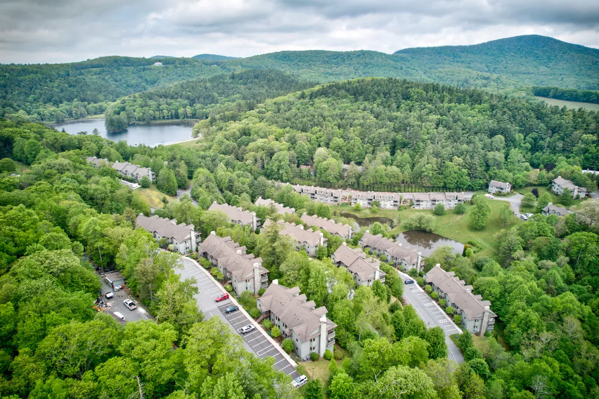 A tranquil scene featuring a serene lake bordered by a lush forest and residential buildings in the background. The scene includes a peaceful waterfront perfect for relaxation, with gentle sunlight illuminating the landscape, inviting guests to unwind by the water.