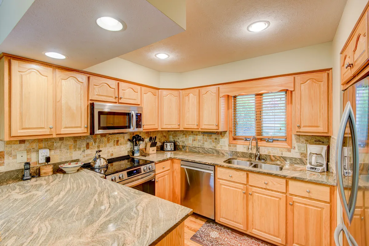 A bright, modern kitchen with a central island, stainless steel appliances, granite countertops, and wood cabinetry. Natural light pours in from a window, highlighting the kitchen’s upscale, functional design.
